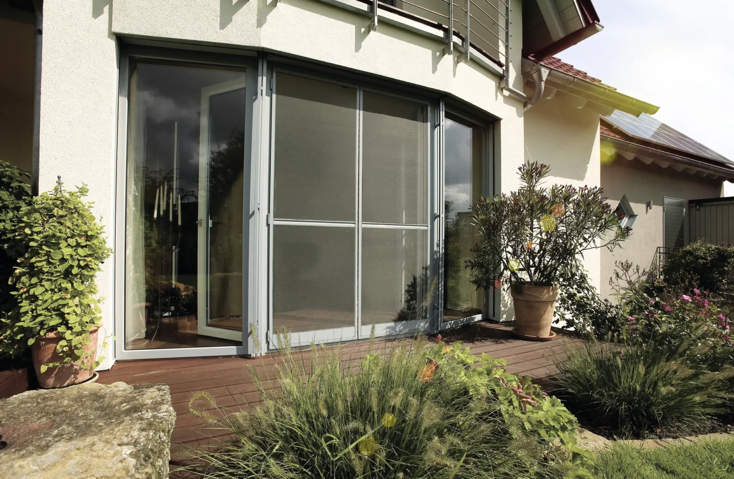 Terrassentür mit Insektenschutzgitter, umgeben von Pflanzen auf einer Holzterrasse an einem Haus